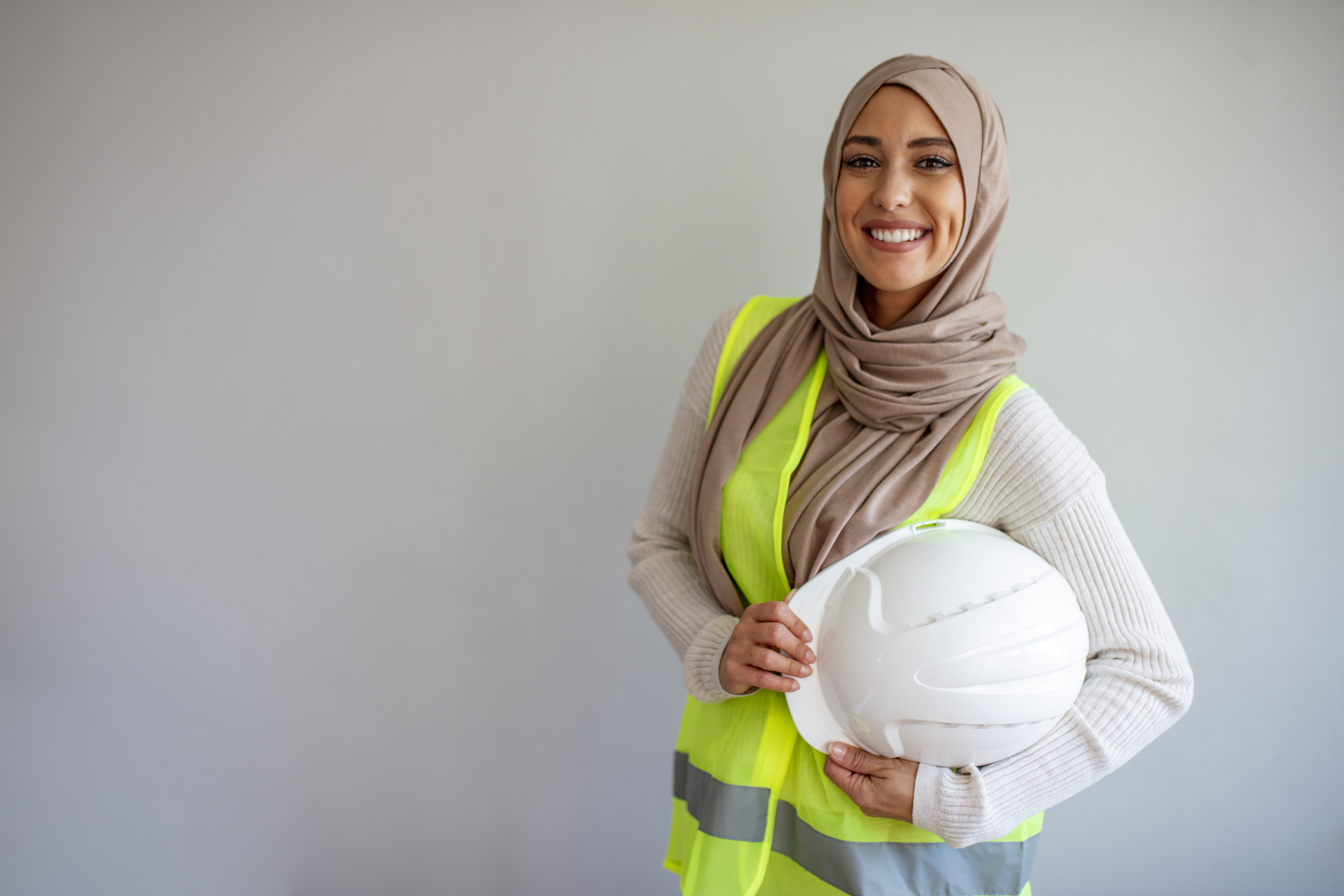 You woman smiling wearing a headscarf, holding a hard hat and wearing a safety vest