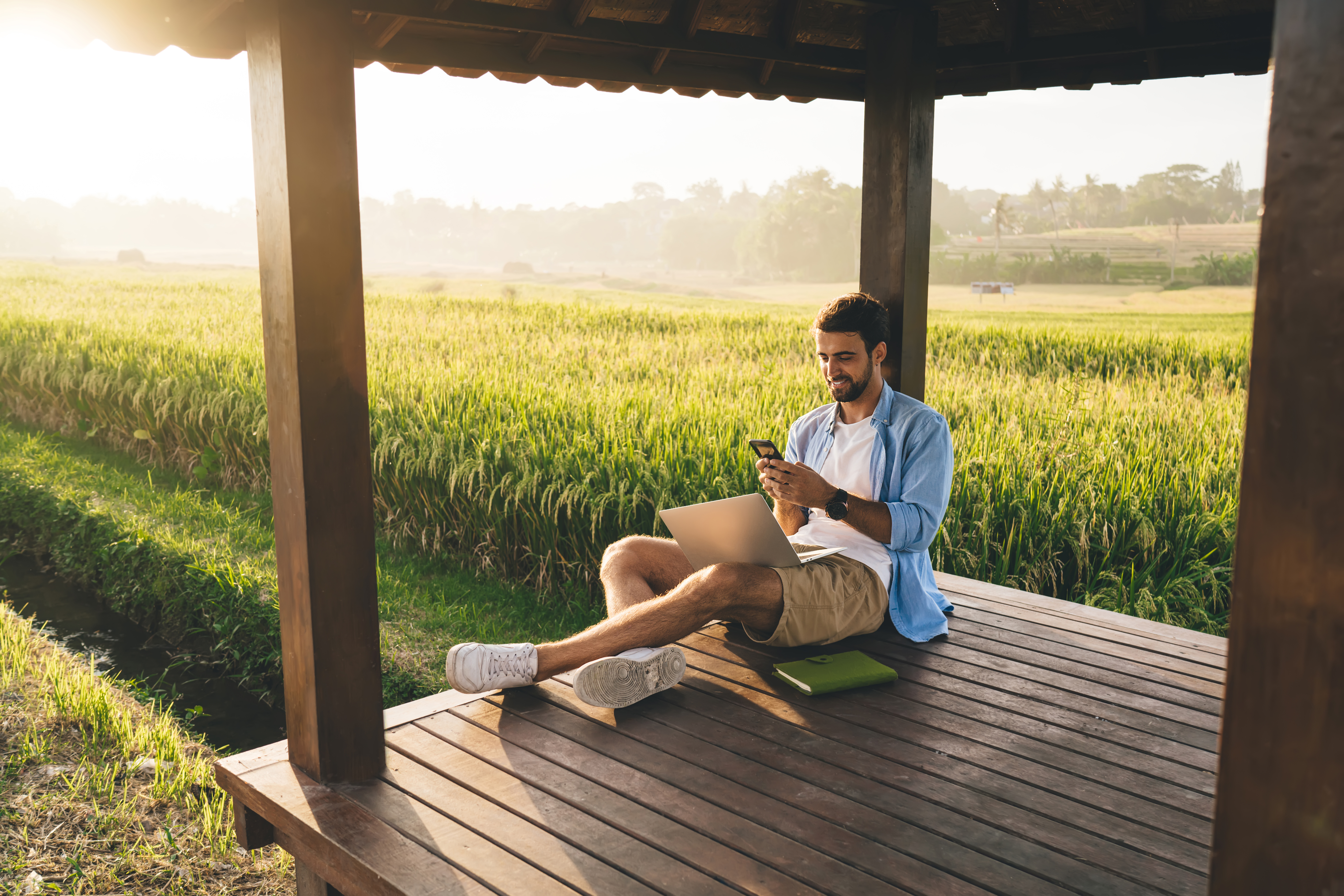 You man on his phone with laptop on lap, sitting in a wooden structure surrounded by cornfield