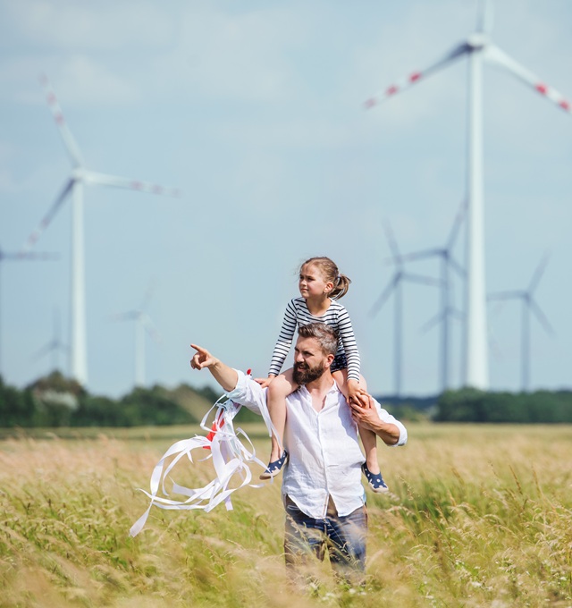 man with girl on his shoulders pointing and looking at something to their right with windmills in the background