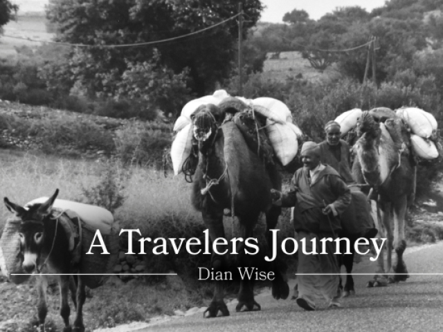 Black-and-white photograph of a rural road with a small donkey in the foreground and two heavily laden camels walking behind it. Two people walk alongside the camels carrying large bundles. Trees, shrubs, and utility lines appear in the background. Overlaid text reads: “A Travelers Journey” and “Dian Wise.”