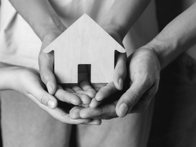 A greyscale image of a family's hands collectively holding a wooden house.