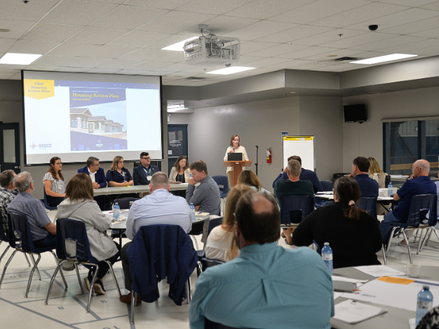 Individuals seated at tables listening to speaker with panel of five people at front of room