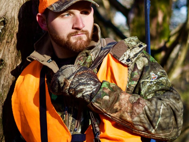 A hunter in camouflage and blaze orange leans against a tree. 