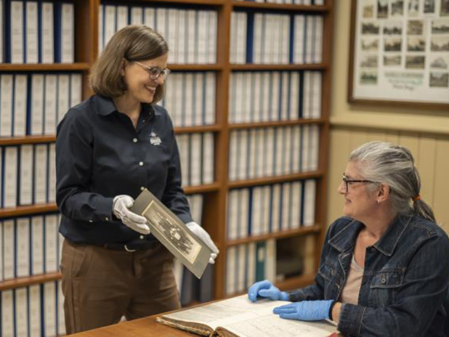 An archivist at the Bruce County Museum and Cultural Centre holds out an artifact to an individual seated with a large book in front of them.