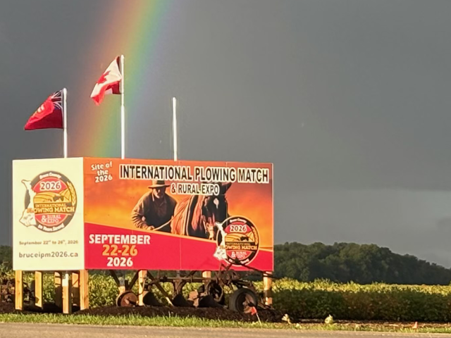 The Bruce County 2026 International Plowing Match and Rural Expo site billboard with flags and a rainbow in the background.