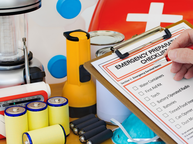 Hand with pen checking off Emergency Preparedness Checklist and emergency prepardness items in background, including flashlight, batteries and canned food.