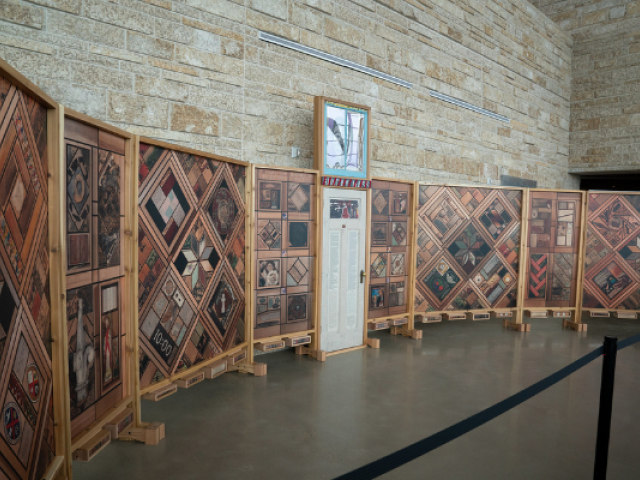 Interior view of The Witness Blanket exhibit at the Bruce County Museum & Cultural Centre. Large wooden panels arranged in a curved display feature hundreds of objects and images collected from residential schools, churches, governments, and Indigenous communities